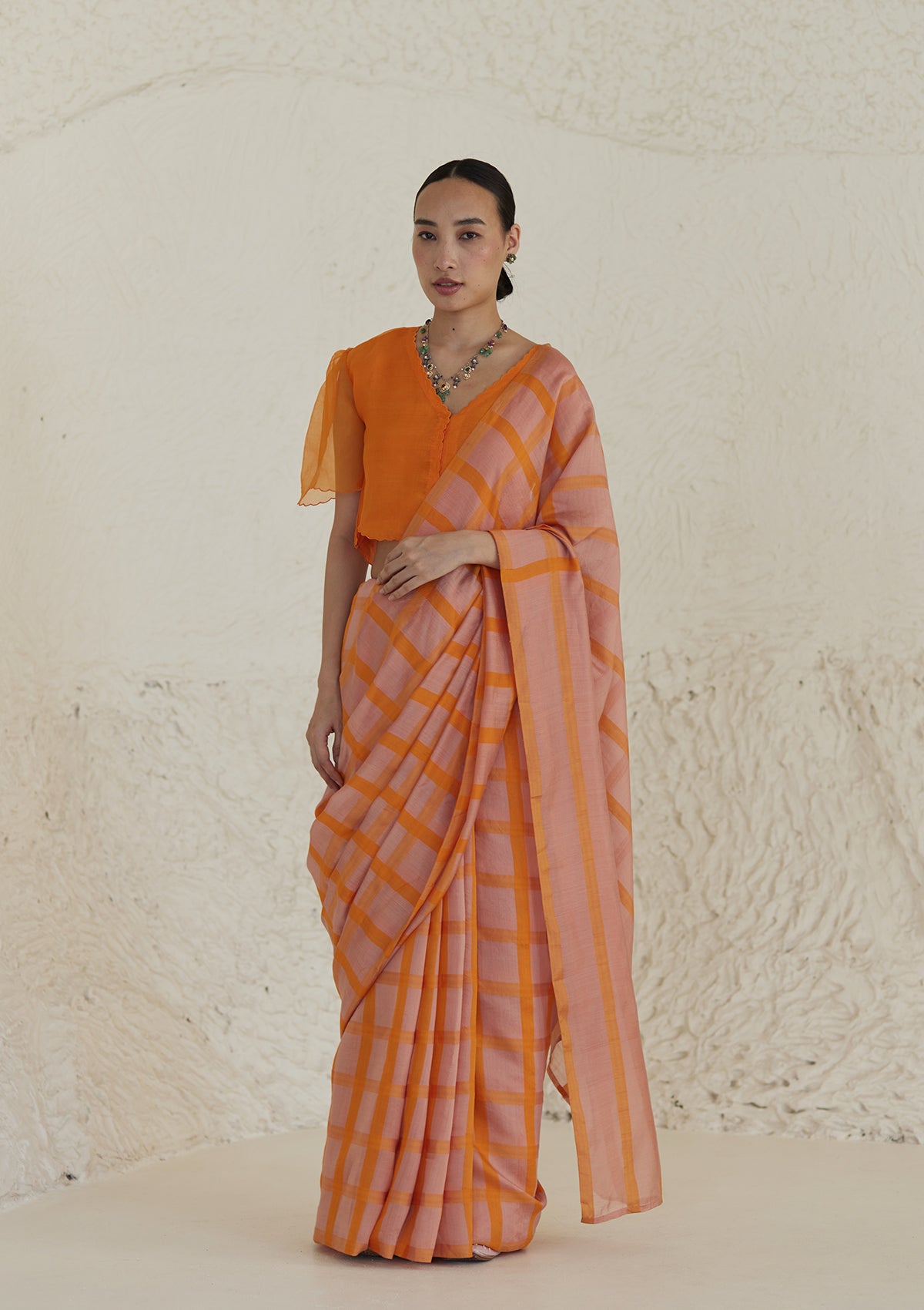 Handloom silk modal saree from Surmaye, having a check pattern with shades of pink and orange. Paired with orange designer blouse, with scalloped embroidery edges all over.