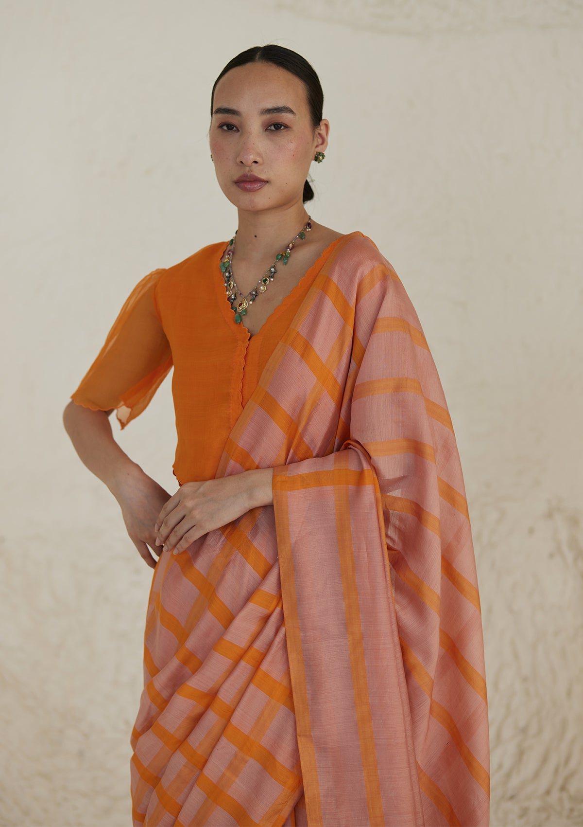 Handloom silk modal saree from Surmaye, having a check pattern with shades of pink and orange. Paired with orange designer blouse, with scalloped embroidery edges all over.